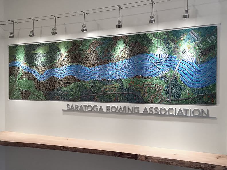 Glass mosaic of a river on a white wall above a wood ledge, lit by cable lights